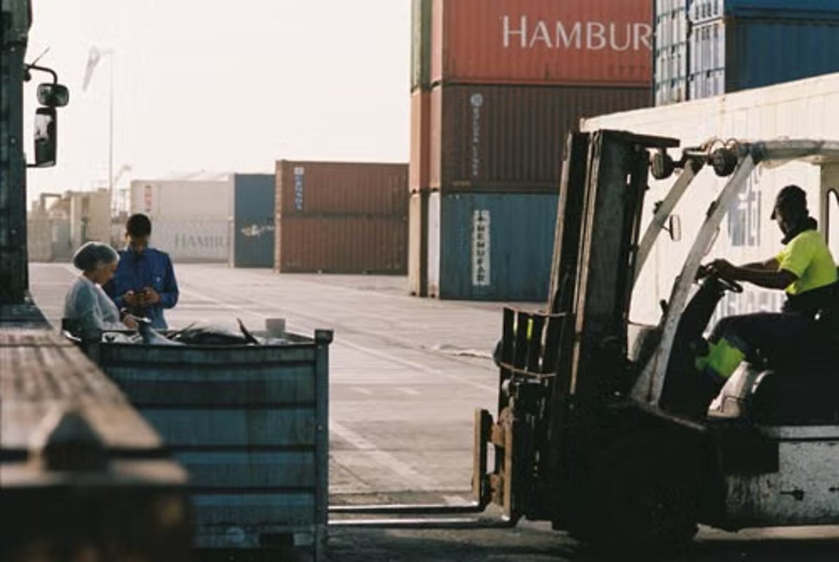 Forklift operating near workers in a port cargo-handling area with shipping containers in the background